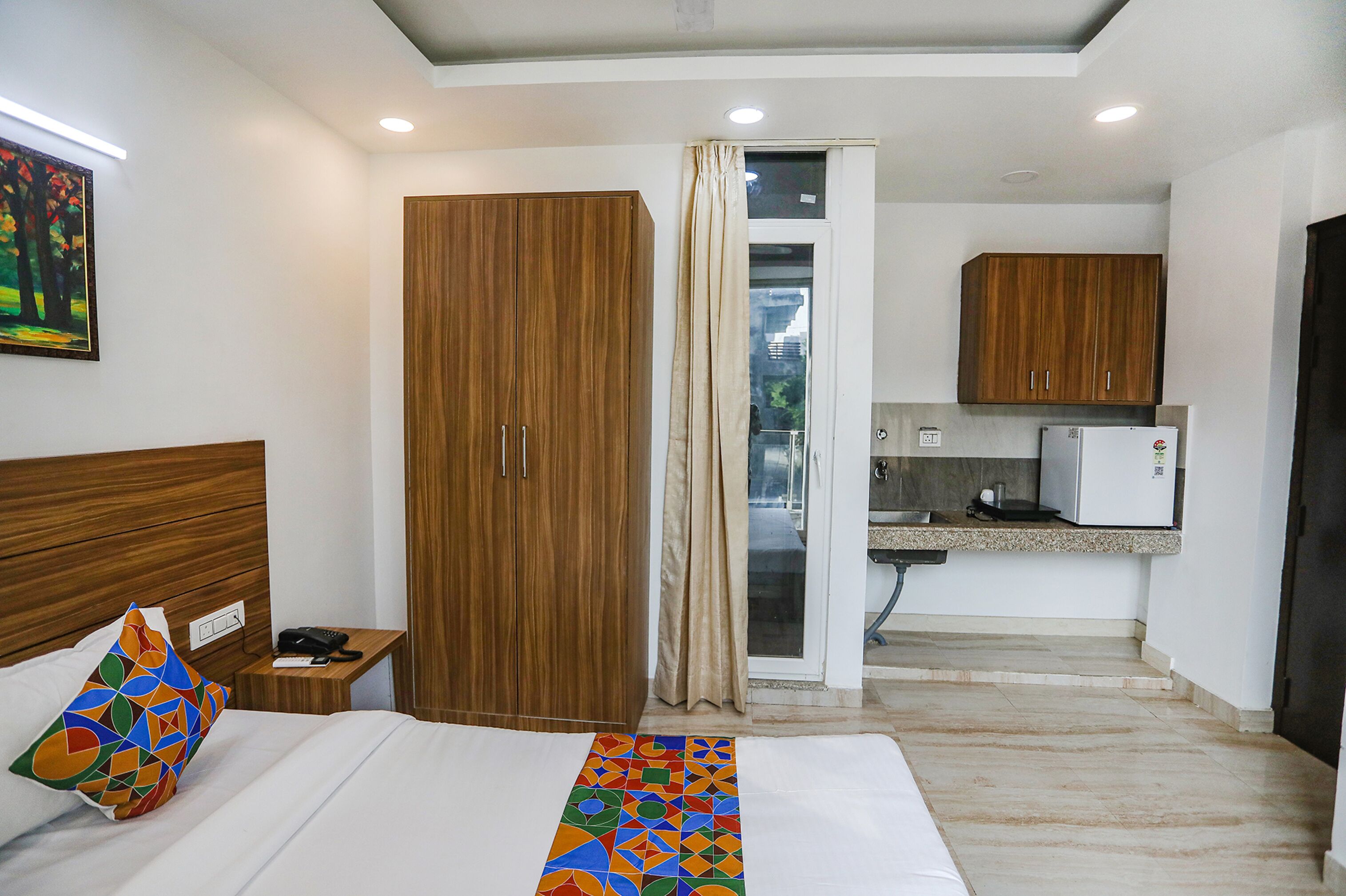 A hotel room with a bed, a nightstand, a wooden wardrobe, a kitchenette, and a bathroom with a glass door.