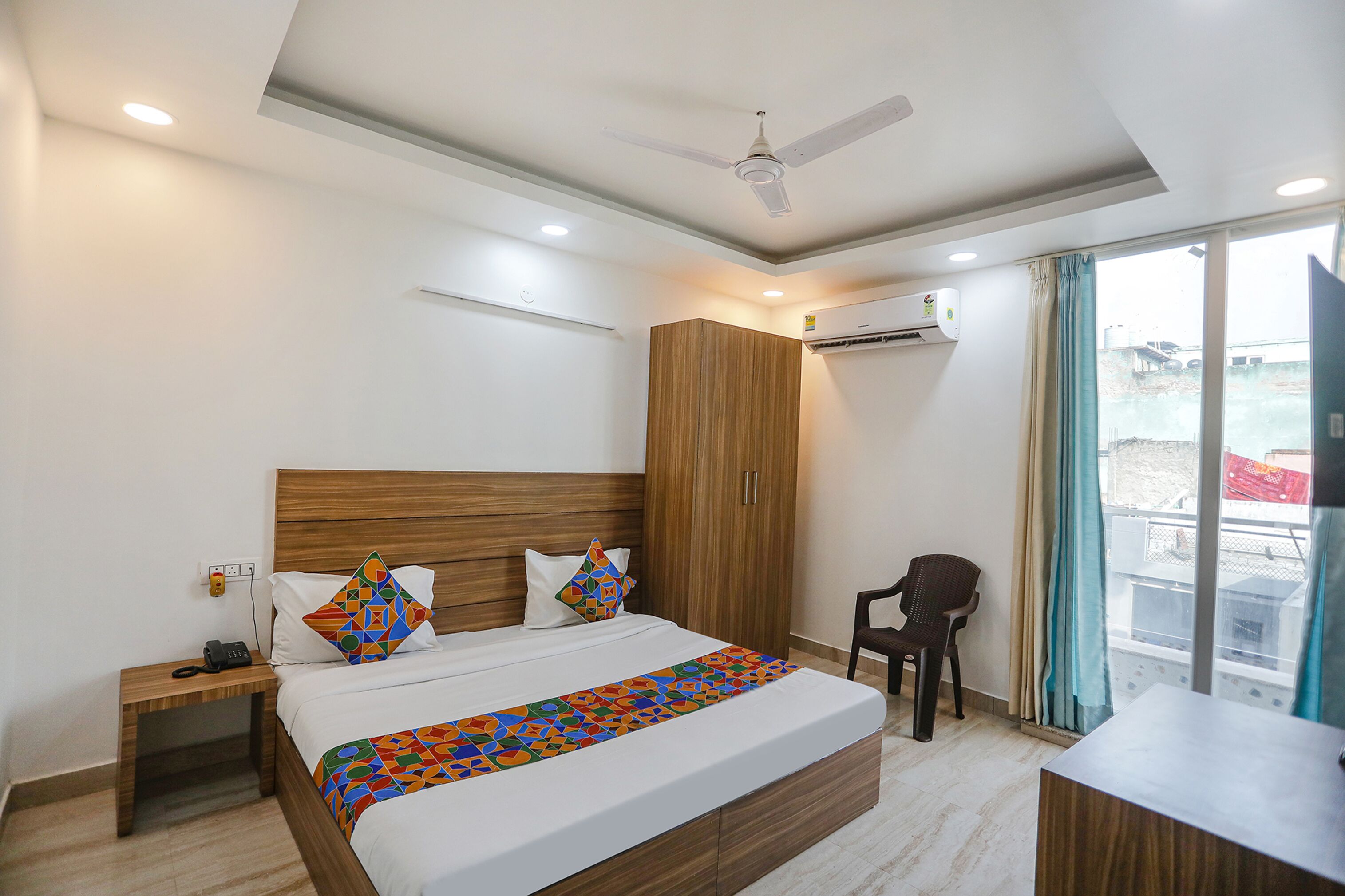 A modern hotel room with a wooden headboard, a bed with patterned bedding, a desk, and a chair.