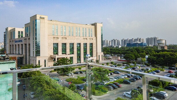 A modern building with a flat roof and large windows, surrounded by a parking lot and greenery. - Fabhotel Unique Stay (Noida)