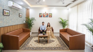 A room with two brown leather sofas, a coffee table, and a patterned rug. There are ceiling fans, an air conditioning unit, and framed artwork on the walls. - Fabhotel Unique Stay (Noida)