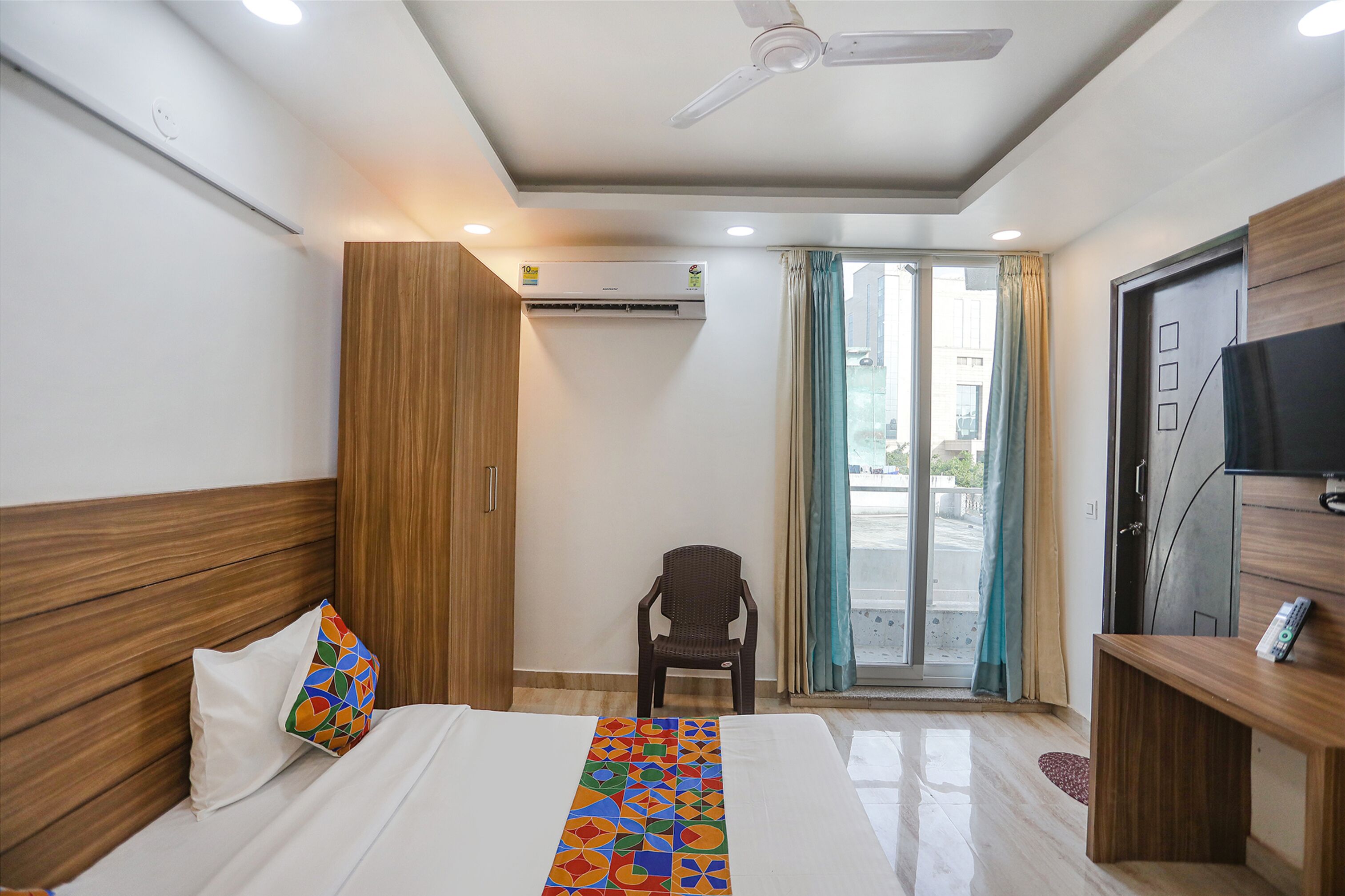 A hotel room with a bed, a chair, a TV, and an air conditioning unit.
