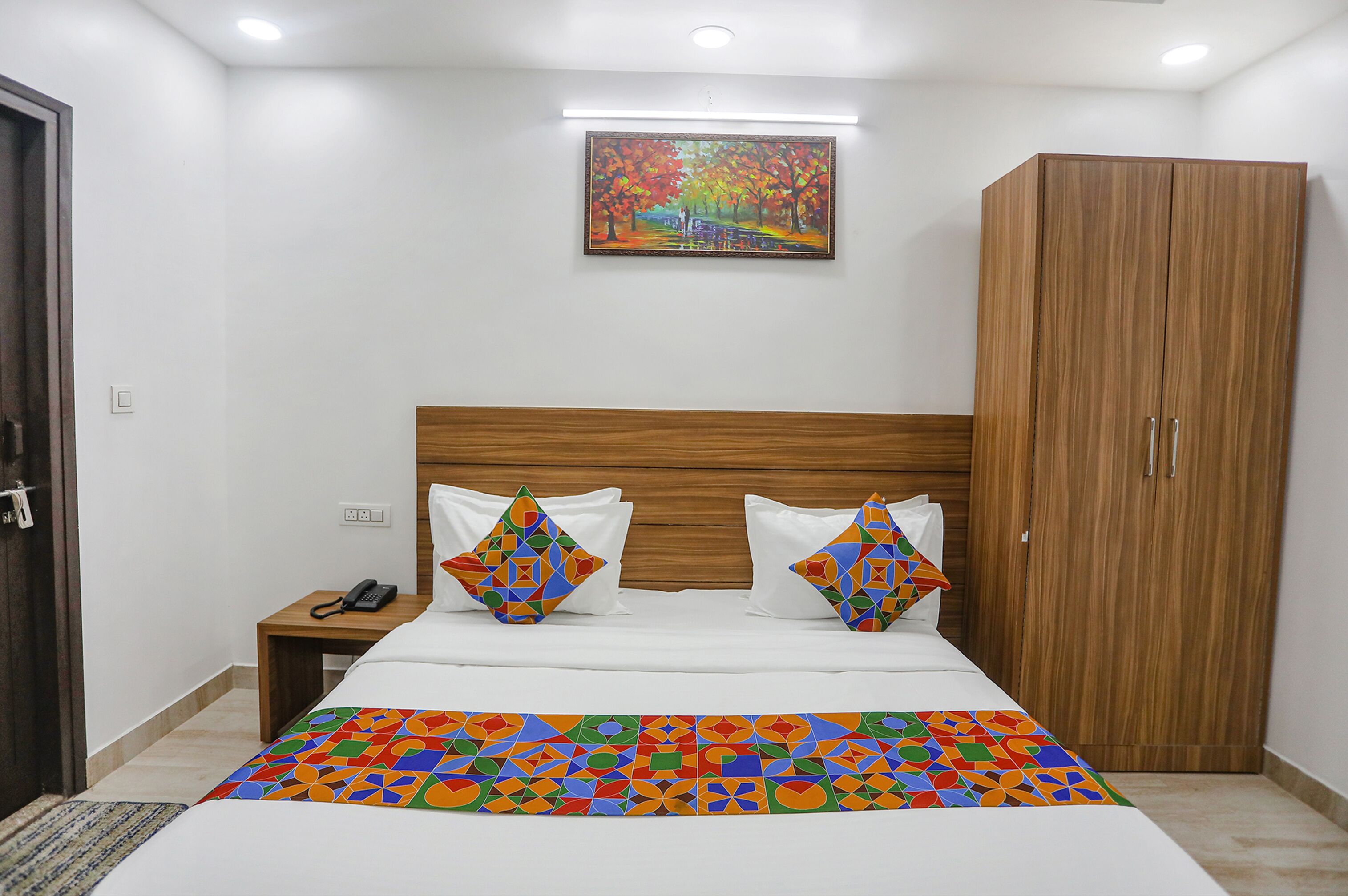 A neatly arranged bedroom with a wooden headboard, a bed with a colorful geometric-patterned bedspread, two decorative pillows, a nightstand with a telephone, and a wardrobe.
