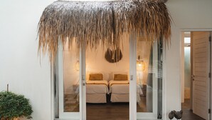 Una habitación con entrada de techo de paja, dos camas con ropa blanca, una lámpara colgante y una pequeña escultura de pájaro en el suelo.