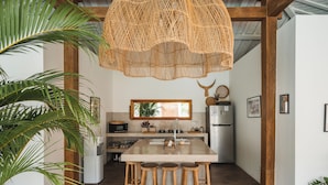 Una cocina con una isla central, taburetes de madera, una lámpara colgante de fibras naturales y una planta grande.
