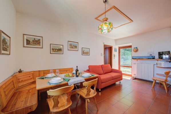 A dining area with a wooden table, chairs, and a red sofa. There are framed pictures on the wall and a kitchen area visible through an open door.