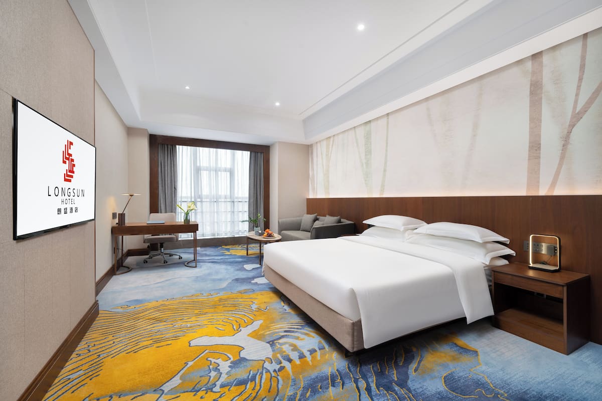 A hotel room with a large bed, a desk with a chair, a small table with a lamp, and a large window with curtains.