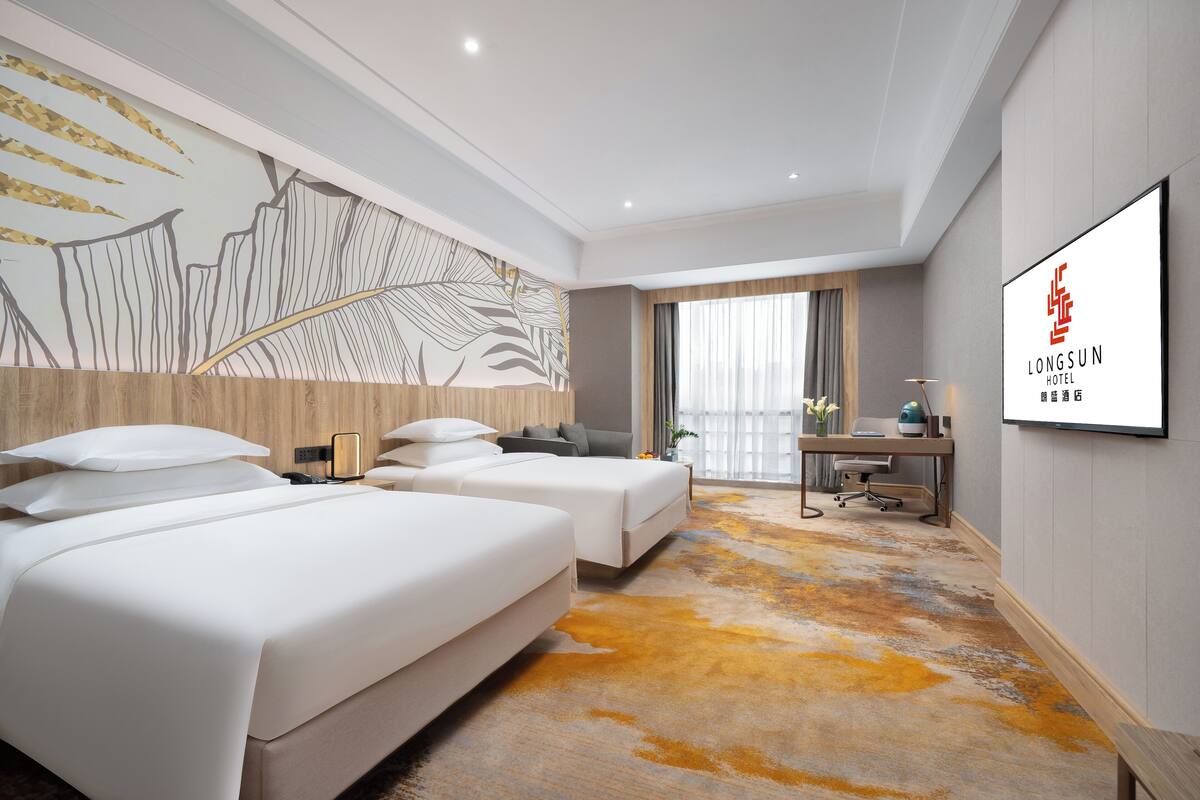 A hotel room with two beds, a desk, a chair, a large window with curtains, and a wall-mounted artwork.