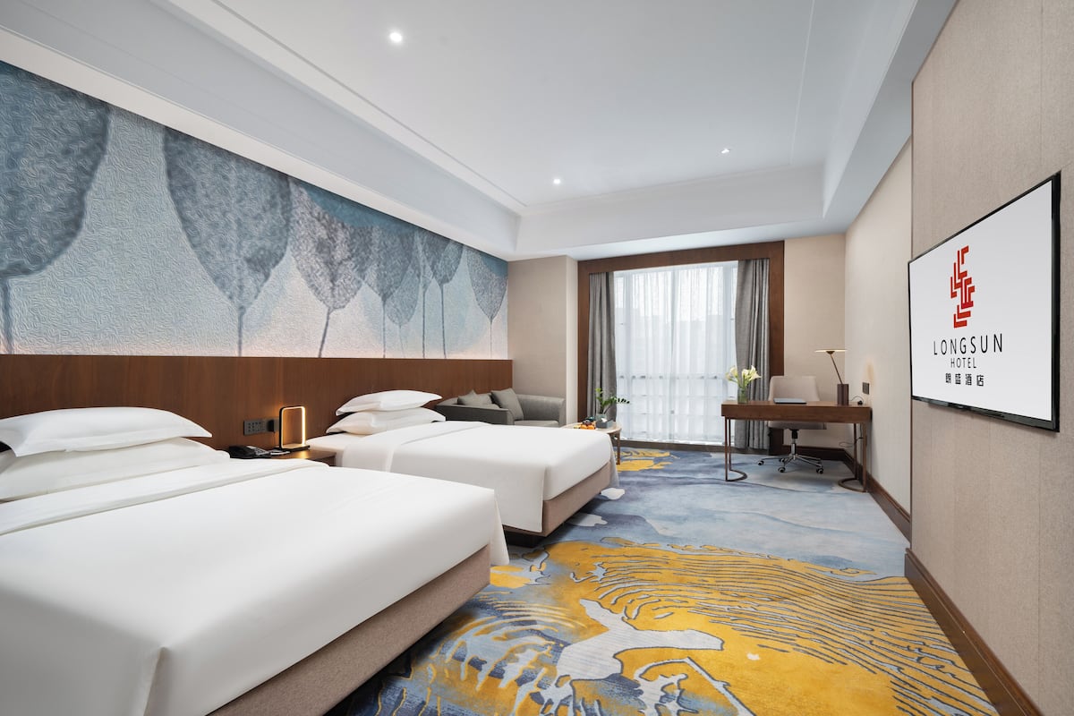 A hotel room with two beds, a desk, a chair, a large window with curtains, and a wall-mounted artwork.