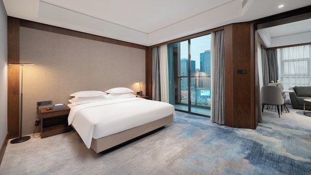 A modern hotel room with a large bed, a desk with a lamp, a view of the city through large windows, and a separate seating area with a table and chairs. - Langson Dongguan - Dalang Songshan Lake (Dongguan)