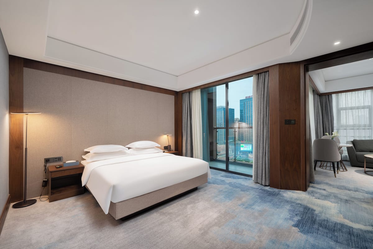 A modern hotel room with a large bed, a desk with a lamp, a view of the city through large windows, and a separate seating area with a table and chairs.