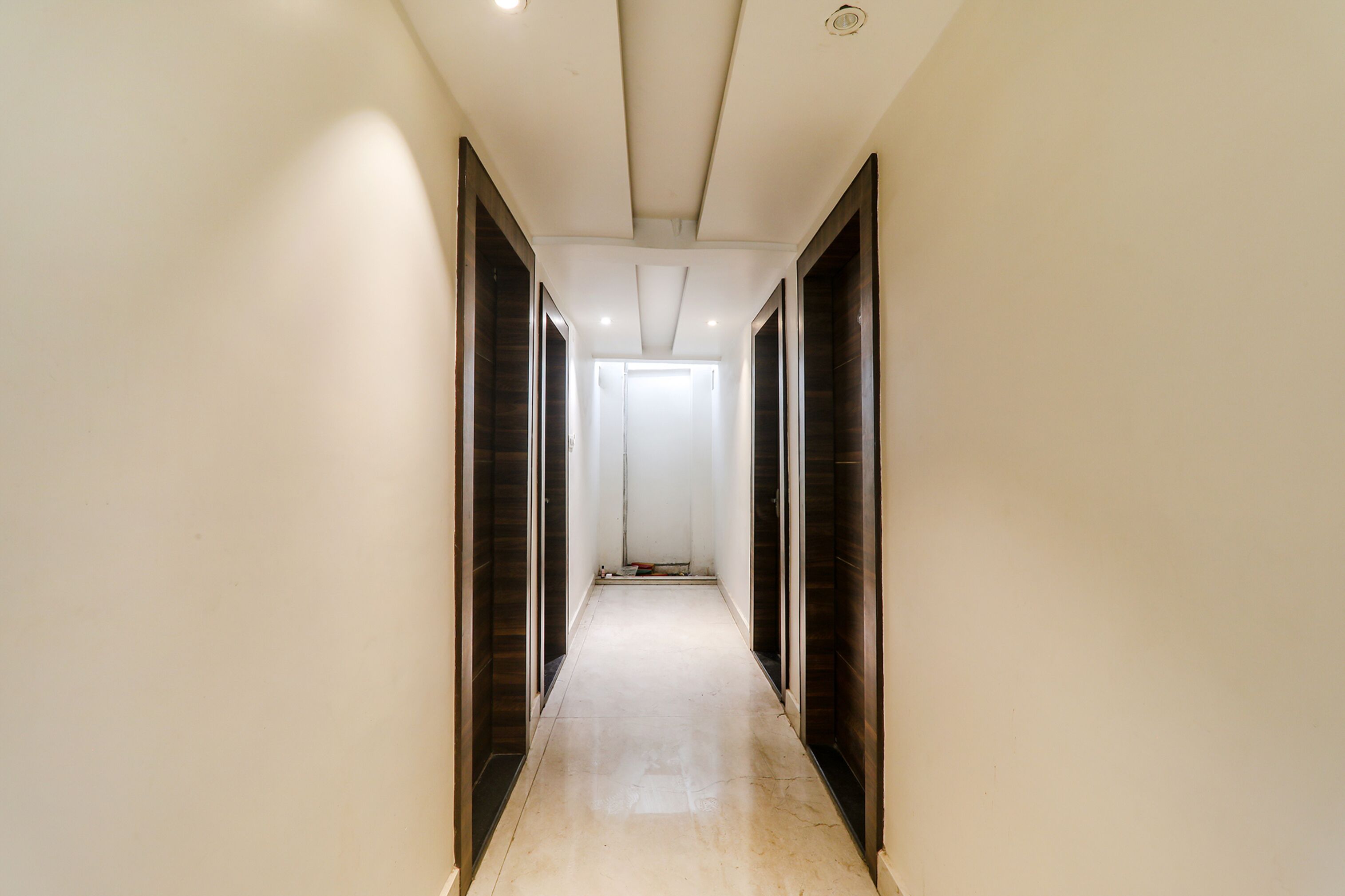 A narrow, well-lit hallway with dark wooden doors on either side and a plain white wall.