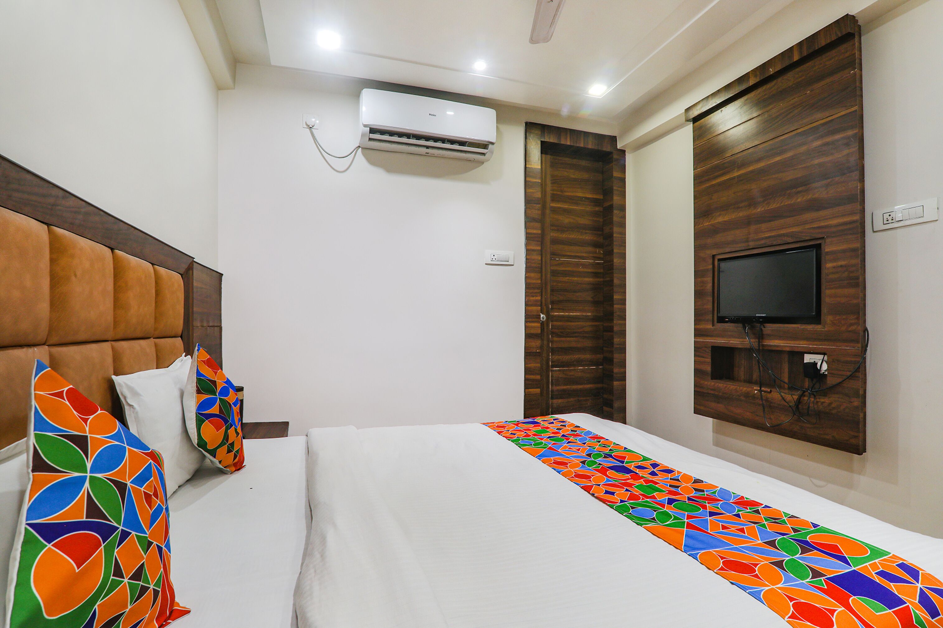 Habitación de hotel con cama, almohadas, equipo de aire acondicionado, televisor montado en un panel de madera y toma de corriente.