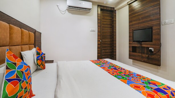 A hotel room with a bed, pillows, an air conditioning unit, a television mounted on a wooden panel, and a power outlet. - Fabhotel Bless Inn (Prayagraj)
