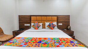 Habitación de hotel con cabecera de madera, colcha con estampado geométrico y colores vivos, dos almohadones decorativos, un sillón de cuero marrón, una mesita de noche con un teléfono y un control remoto, y un equipo de aire acondicionado instalado en la pared.