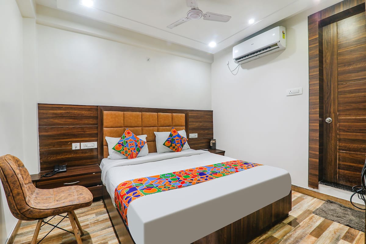 A bedroom with a wooden headboard, a bed with colorful pillows, a chair, a bedside table, and an air conditioning unit.
