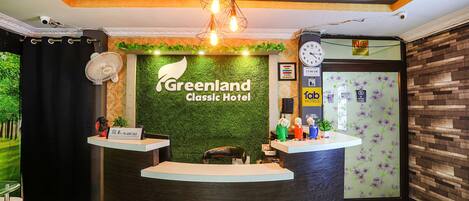 Un comptoir d’accueil à l’hôtel Greenland Classic, agrémenté d’un mur vert, d’une horloge et d’un ventilateur.