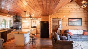 Een gezellig chaletinterieur met houten details, een keuken met een achterwand van steen, een eethoek en een woonkamer met een bank en vloerkleed.