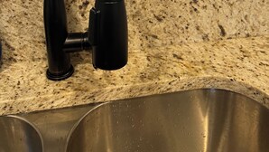Eine Küche mit einem schwarzen Wasserhahn, einer Arbeitsplatte aus Granit, einem Edelstahl-Spülbecken und einem flachbildschirmfernseher an der Wand.