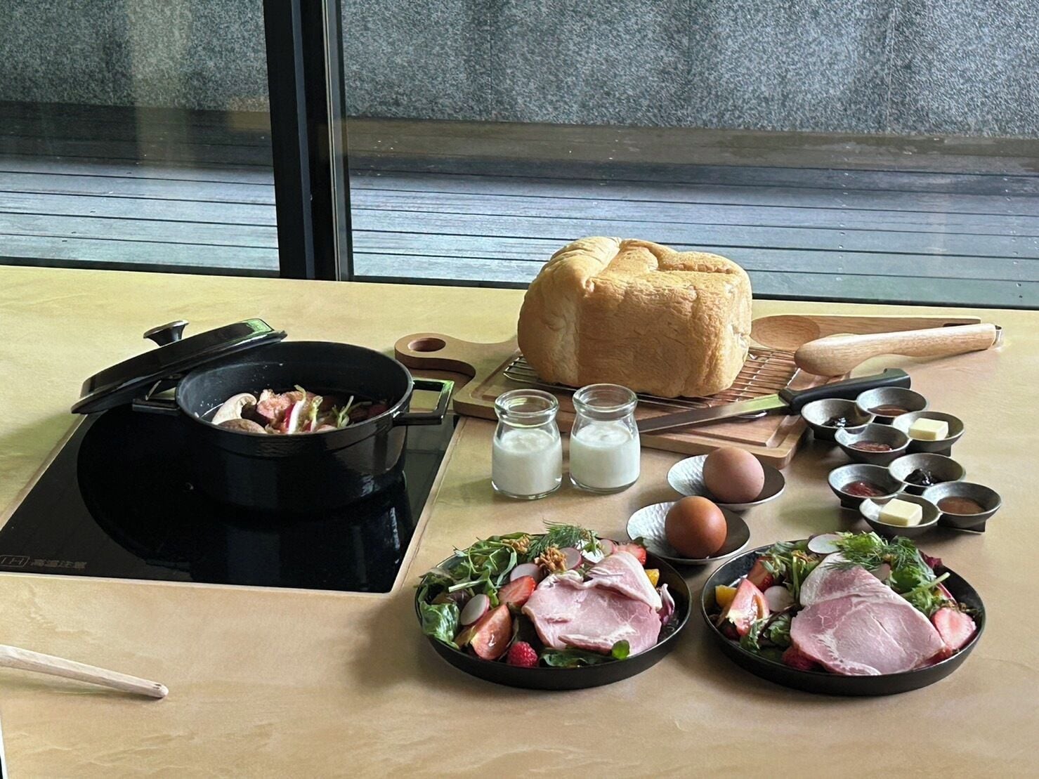 A breakfast setup with a bread roll, a pot of food, milk, eggs, and various condiments on a table.