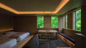 Ein modernes Hotelzimmer mit Bett, Sofa, kleinem Tisch und Schreibtisch. Große Fenster bieten Blick auf eine grüne Umgebung.