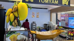 Un étal de nourriture avec une grande enseigne indiquant « DELI BANGKOK TOMYAM » et une variété de fruits frais exposés, comme des bananes et des mangues.