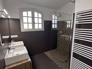 Et moderne badeværelse med mørke fliser, et hvidt møbel med tre håndvaske, et bruseområde med glasdør og en håndklædestang.