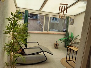A room with a large window, a bamboo plant, a black chair, a woven chair, and a small table.