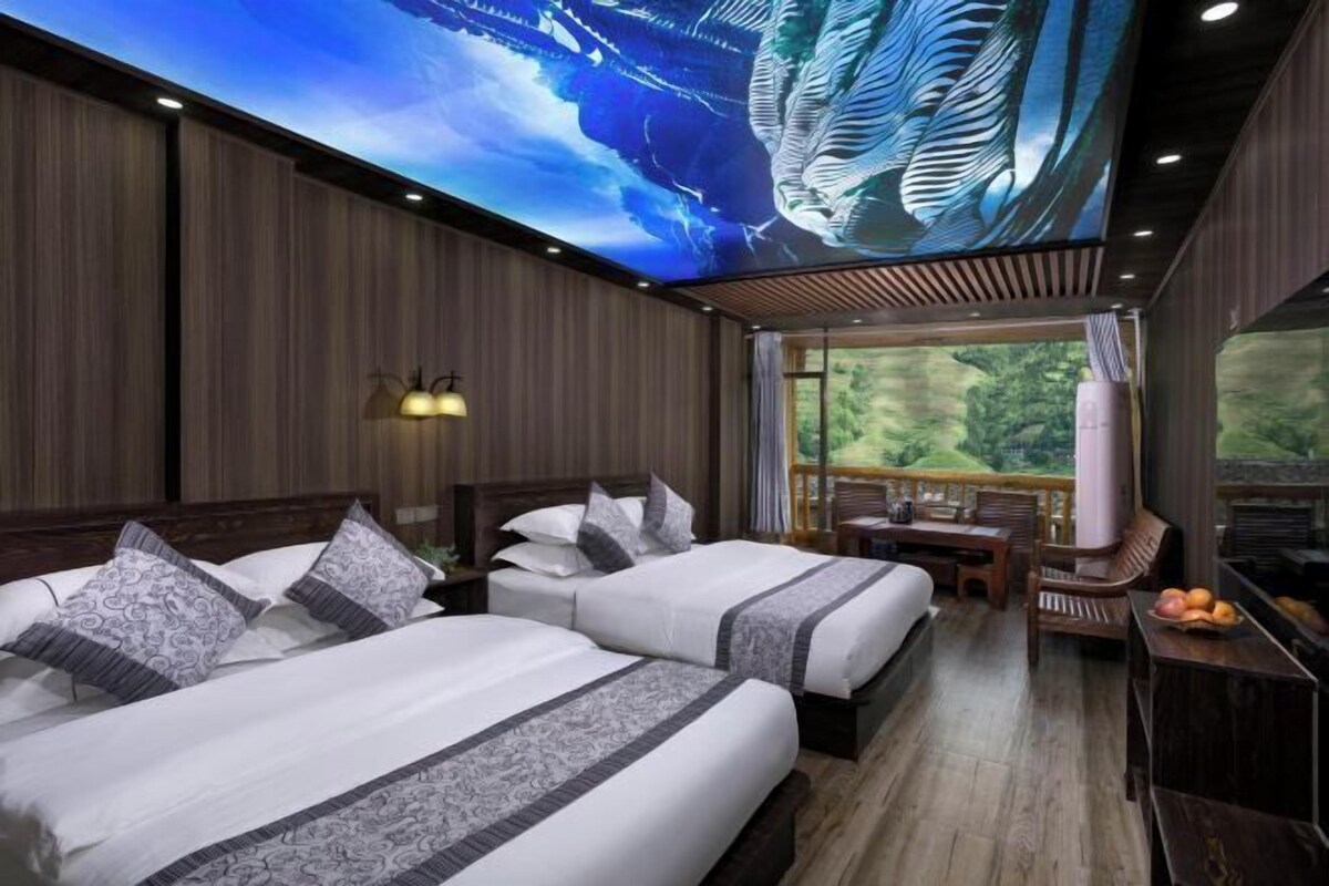 A hotel room with two beds, a TV screen on the ceiling, a balcony with a view, and a small table with chairs.