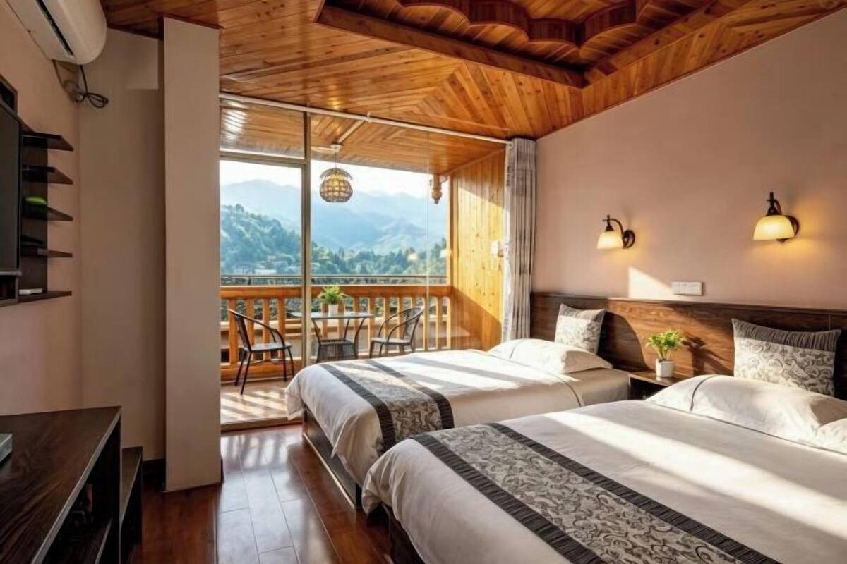 Ein Zimmer mit zwei Betten, einer Holzbalkendecke, einem Balkon mit Bergblick und einem Tisch mit Stühlen.