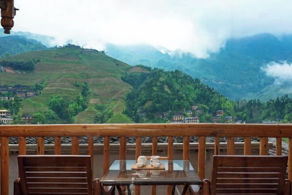 A wooden balcony with a table and chairs, overlooking a mountainous landscape with terraced fields and buildings.