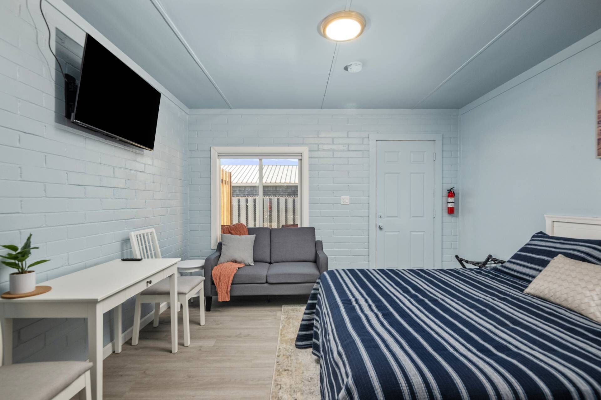 Une chambre à coucher avec un lit, un bureau, un canapé gris, une fenêtre et une porte.