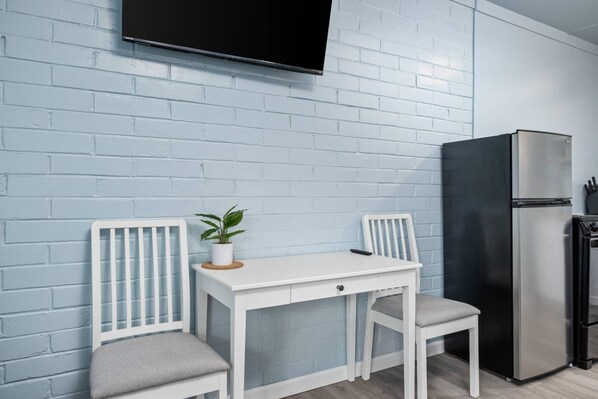 Una cocina compacta con una mesa y sillas blancas, un refrigerador negro y un televisor montado en una pared de ladrillo azul claro.