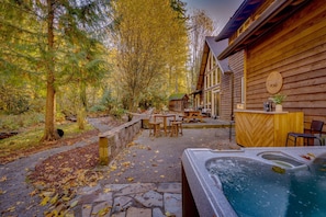 A wooden cabin with a hot tub, outdoor seating, and a stone pathway surrounded by trees.