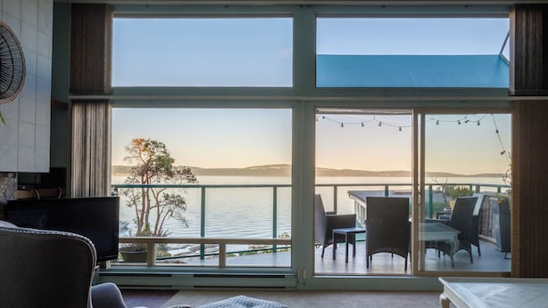 A spacious living area with a large window overlooking a body of water, furnished with a sofa, chairs, and a small table. - INN OF THE SEA - Stunning views~ Oceanfront Pool & Hot Tub, Private Dock (Ladysmith)
