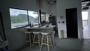 A modern kitchen with a large window overlooking a scenic landscape, a bar area with stools, and a ceiling fan. - Ocean/Beachfront Living ~Rustic Fishing Village w/Upscale Amenities HAVE IT ALL! (El Gigante (Playa Gigante), Tola)