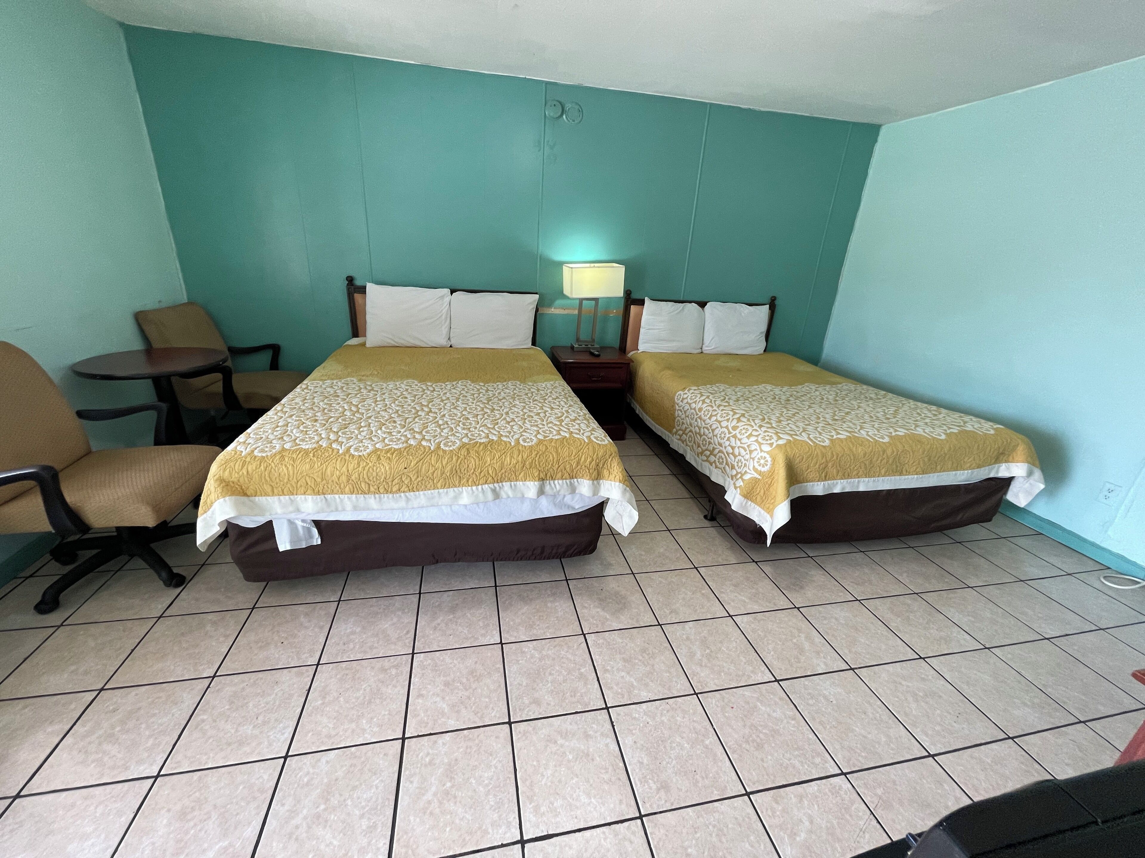 A hotel room with two beds, a small table, a chair, and a nightstand with a lamp.