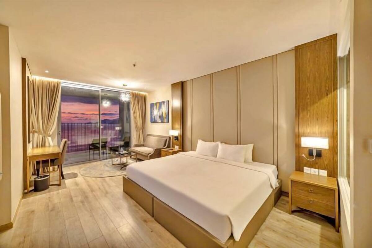 A hotel room with a large bed, a desk, a chair, a nightstand, and a view of the sunset through a glass door.