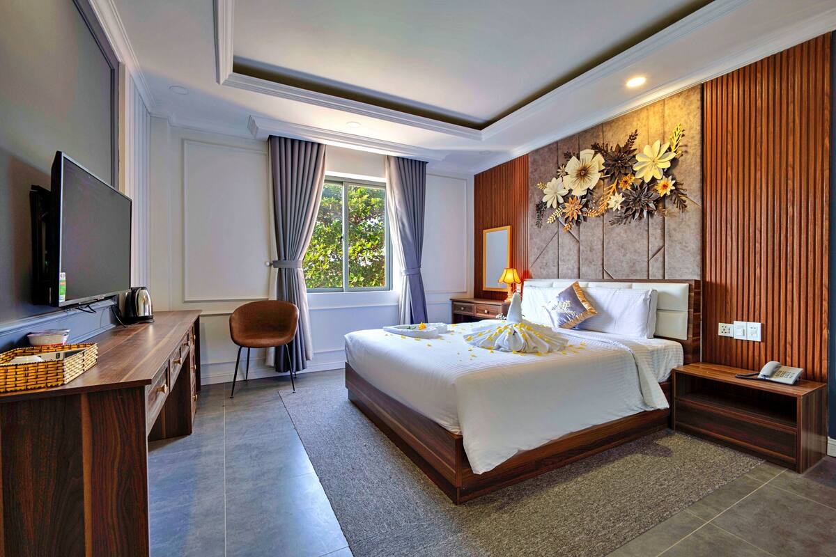 A modern hotel room with a large bed, a desk with a flat-screen TV, a chair, a bedside table with a lamp, and a decorative wall panel.