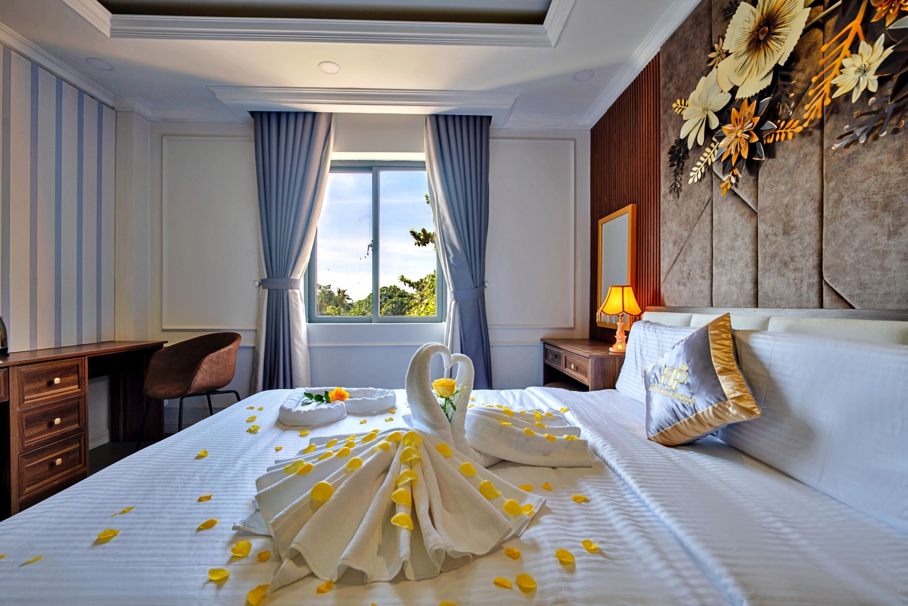 A hotel room with a bed, a desk, a chair, a nightstand with a lamp, and a window with curtains.