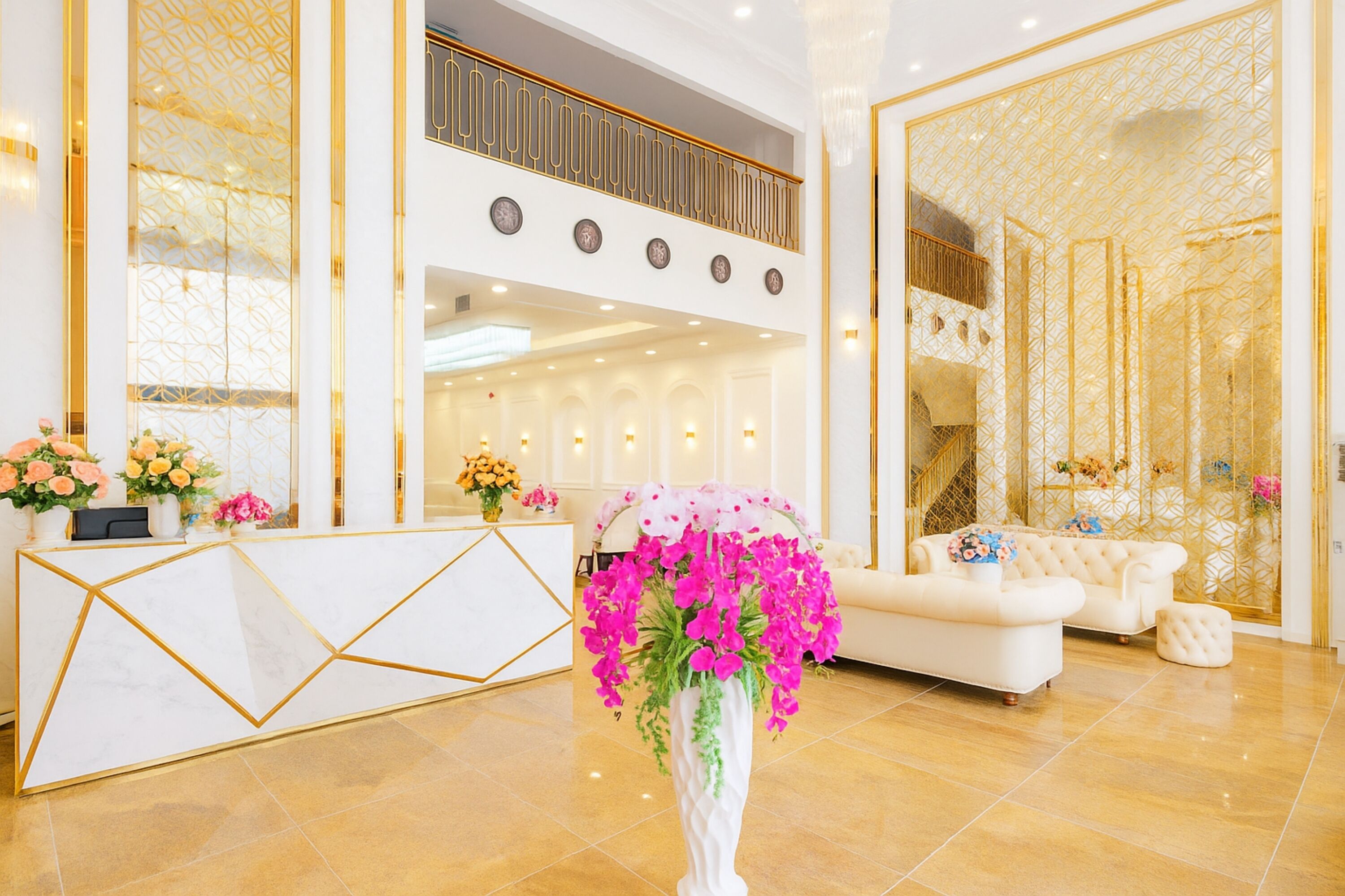Un área de recepción con un mostrador blanco, un jarrón grande con flores rosas y un sofá blanco con marco dorado.