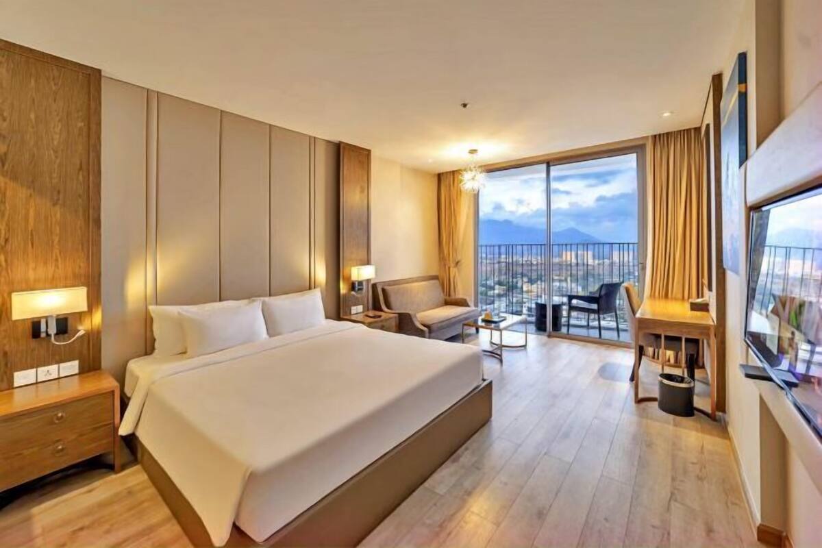A hotel room with a large bed, a desk, a chair, a small table, and a balcony with a view.