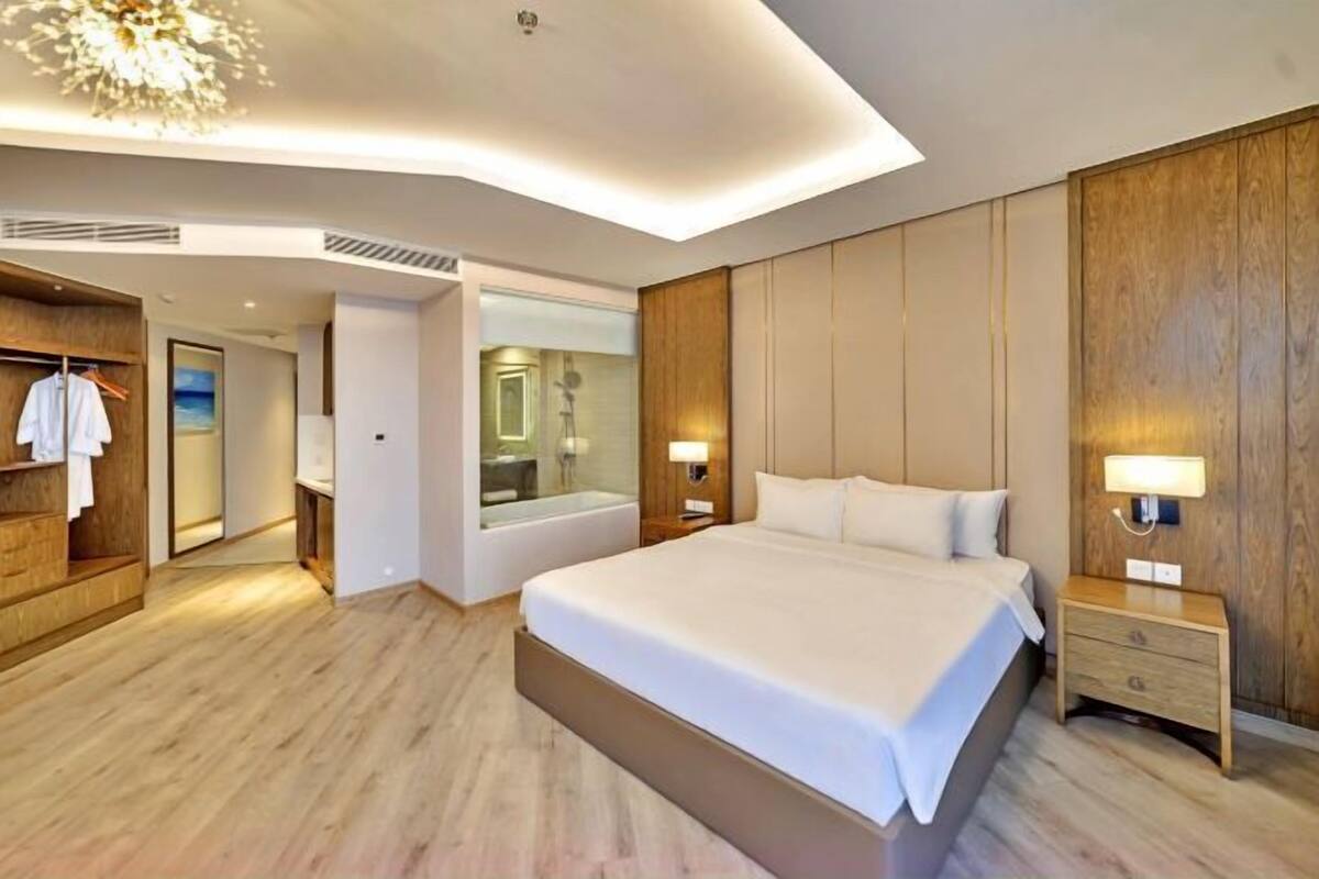 A modern hotel room with a large bed, wooden panel walls, a walk-in closet, and a bathroom with a glass shower.