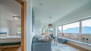 Un dormitorio moderno con piso de madera, una cama, un sofá gris y un ventanal con vistas a la montaña.