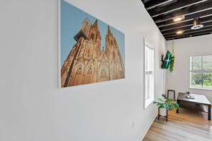 Une pièce avec un grand tableau représentant une cathédrale, une fenêtre avec des stores, un bureau et une chaise.