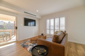 A modern living room with a brown leather sofa, a round black coffee table with a book, a TV mounted on the wall, and a large window with blinds.