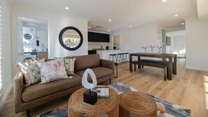 A modern living room with a brown sofa, a round mirror, and a dining area with a wooden table and benches. - Getaway on Curran. EV + Firepit. Walk to CBD. (Orange)