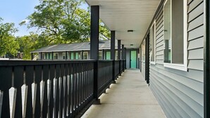 A covered corridor with a black railing, leading to a building with a green door.