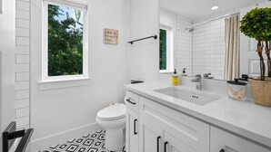 A modern bathroom with a white sink, toilet, and vanity. There's a window with a view of trees, a mirror, and a sign on the wall. - Cherrywood Home Near UT / ATX / Private Backyard (Austin)