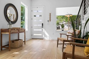A room with a wooden floor, a white door with frosted glass panels, a console table with baskets, a chair with a cushion, and a view of greenery outside.