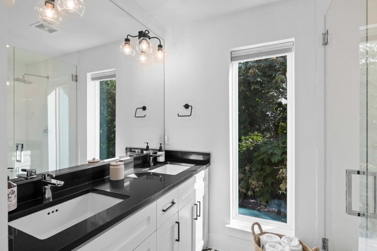 A modern bathroom with a black countertop, two sinks, and a large mirror. There is a window providing a view of greenery outside.